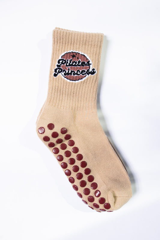 Single tan Pilates sock from Dew Health & Wellness laid flat to show rubberized grips and “Pilates Princess” logo embroidery.