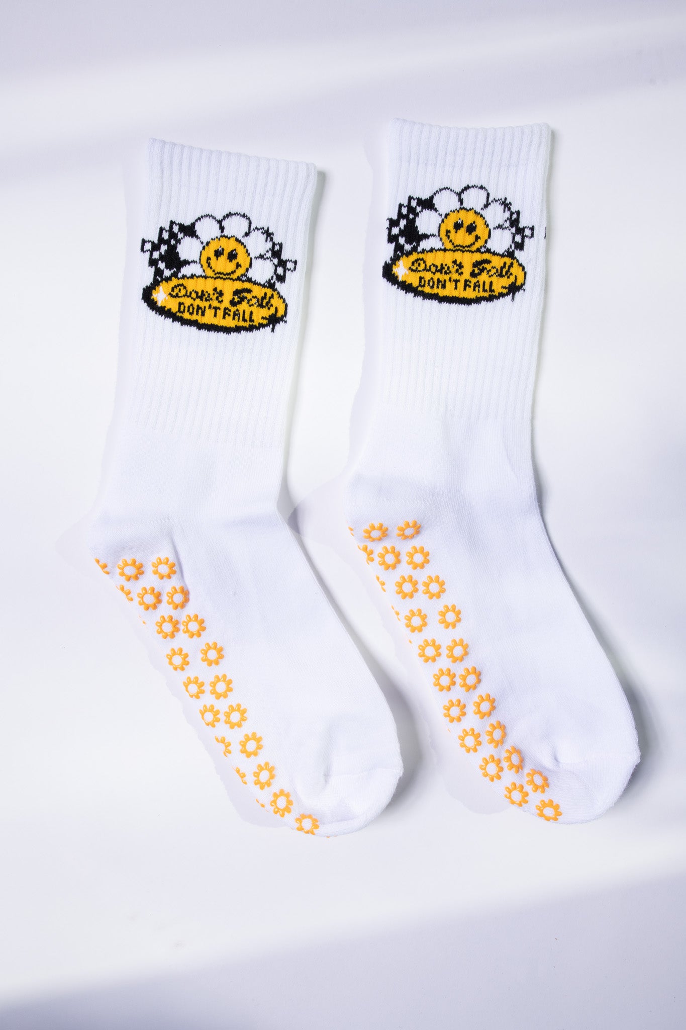 Flat lay of white grip socks from Dew Health & Wellness, featuring daisy-patterned soles and a cheerful “Don’t Fall” daisy patch.