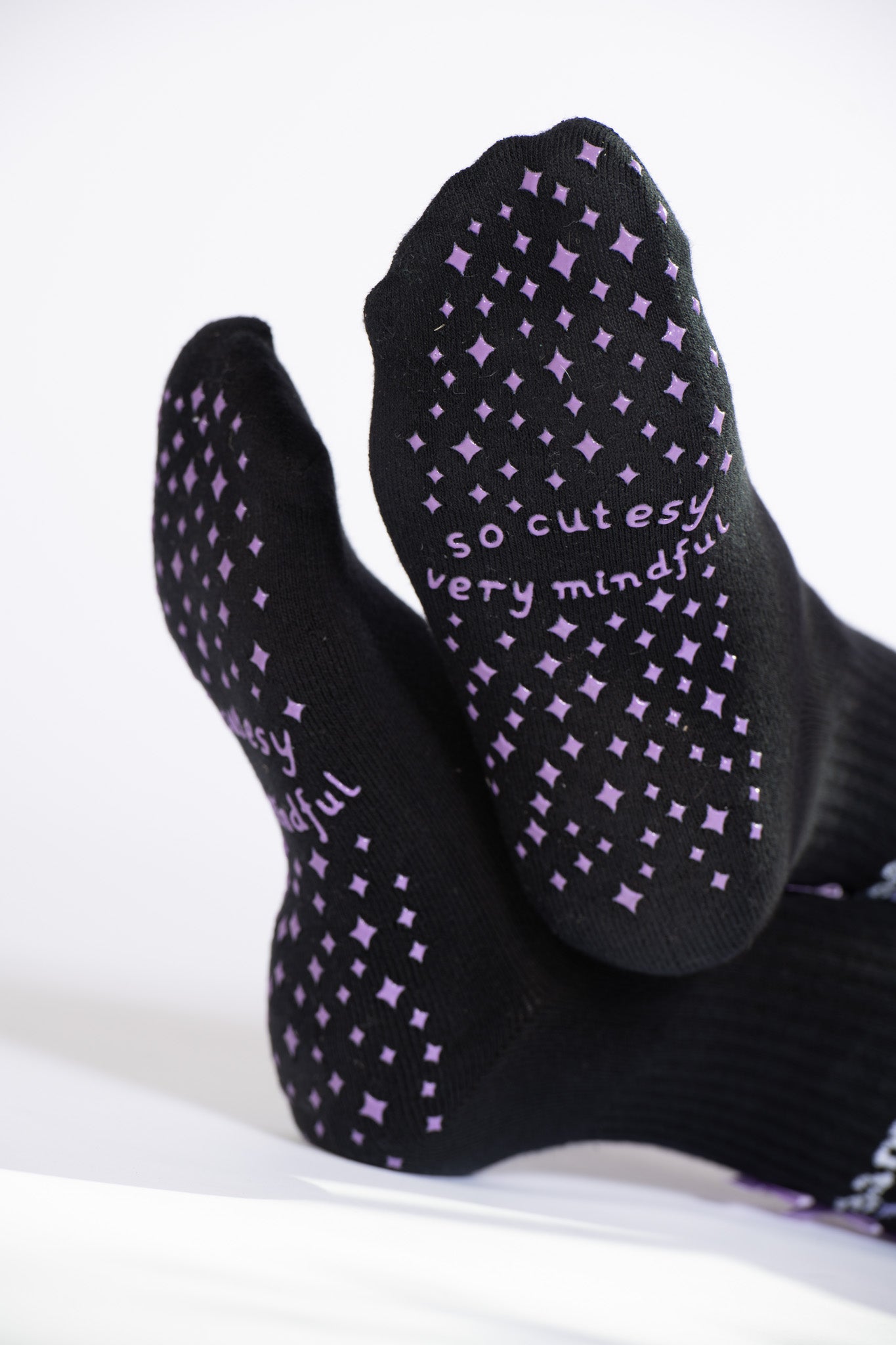 Close-up of lavender grip pattern and “so cutesy very mindful” text on soles of black Pilates socks.