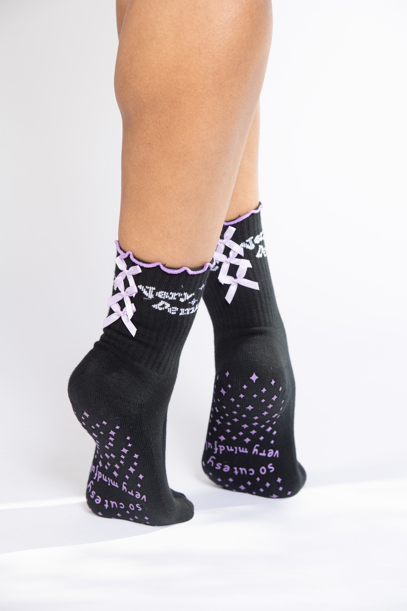 Model on tiptoe wearing black grip socks with lavender bows and text “so cutesy very mindful” on soles.