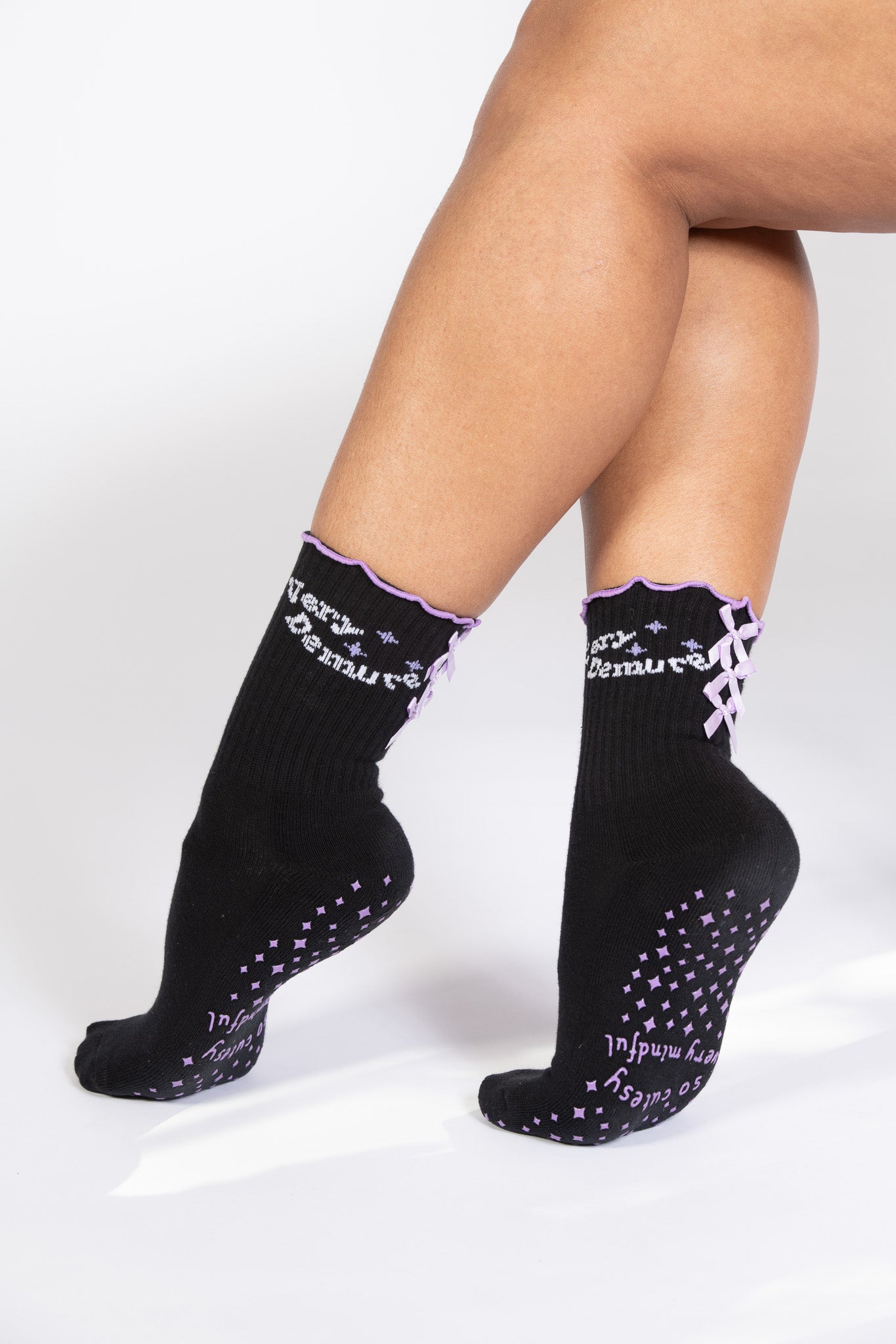 Model wearing black Pilates grip socks with lavender bows, frilly edges, and lavender grip pattern.