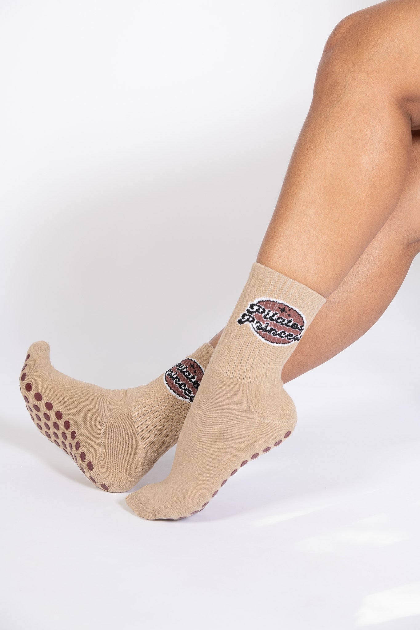 Model wearing Dew Health & Wellness Pilates Princess grip socks in tan with brown grips, posed on tiptoe to highlight functionality.