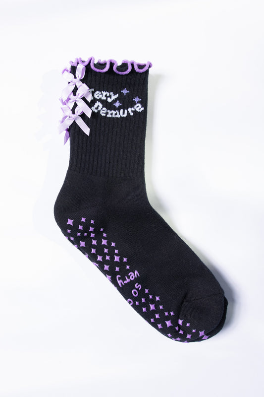 Flat lay of black Pilates grip sock with lavender bows, ruffled trim, and star-pattern lavender grips.