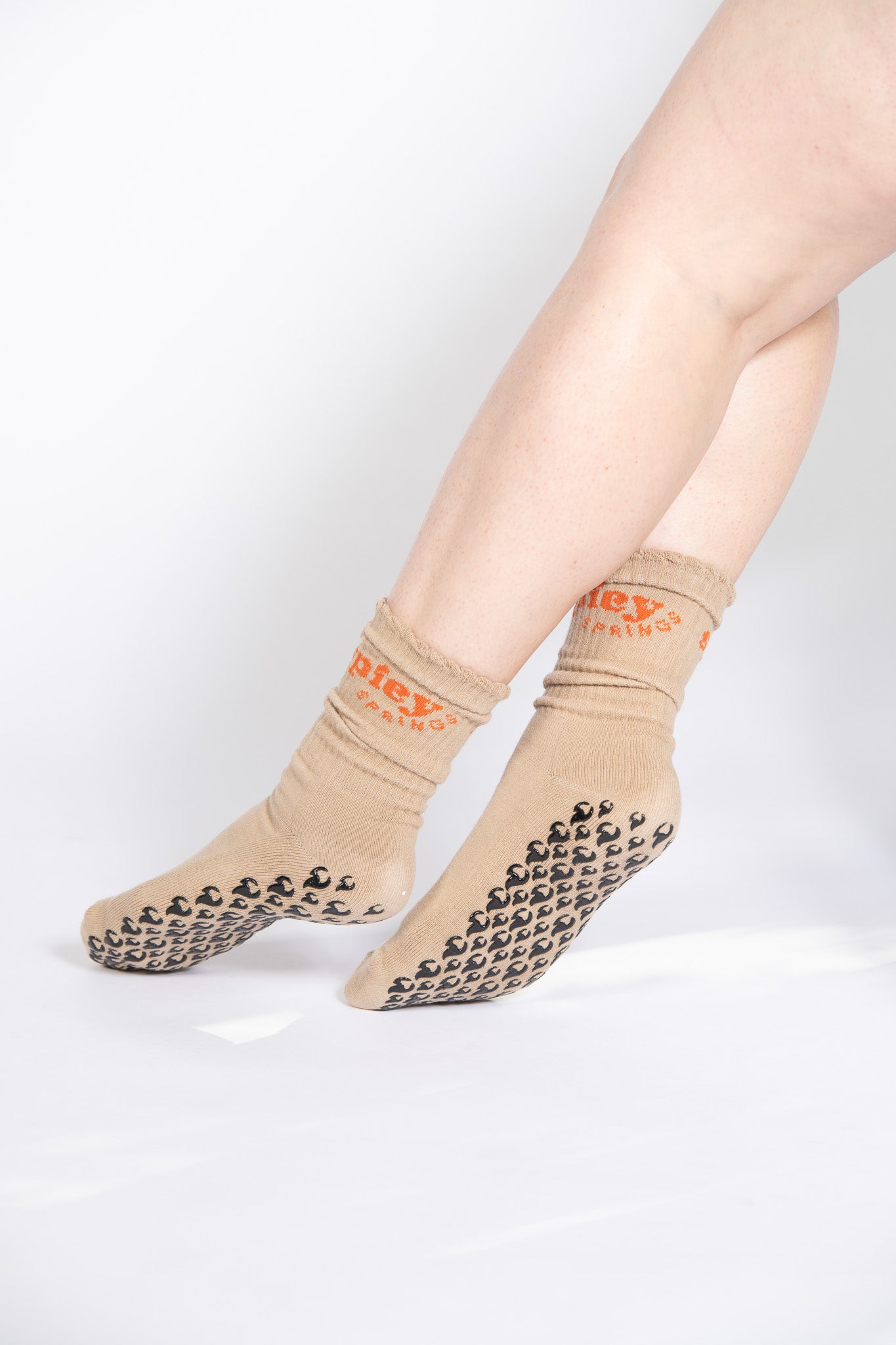 Model wearing tan grip socks with black flame tread and orange embroidered cuff text, posed mid-step.