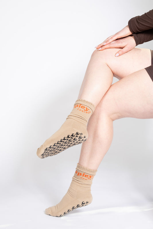 Model seated in tan Pilates socks with black flame-shaped grips and orange “spicy springs” cuff detail.