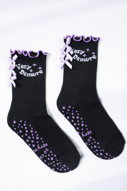 Flat lay of black Pilates socks with lavender text, bows, and non-slip diamond grip pattern.