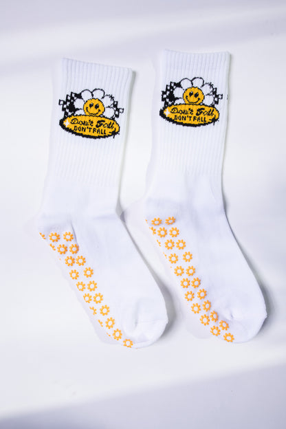 Flat lay of white grip socks from Dew Health & Wellness, featuring daisy-patterned soles and a cheerful “Don’t Fall” daisy patch.