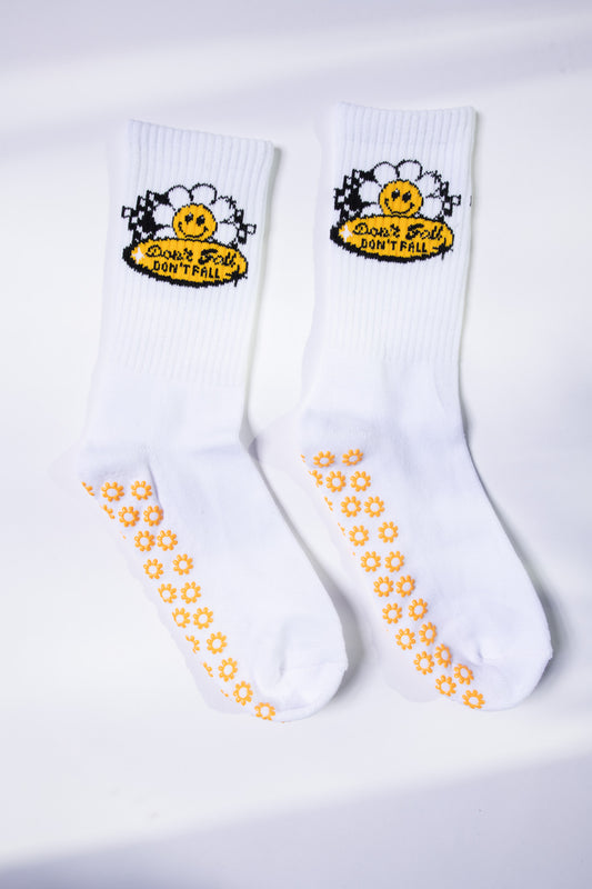 Flat lay of white grip socks from Dew Health & Wellness, featuring daisy-patterned soles and a cheerful “Don’t Fall” daisy patch.