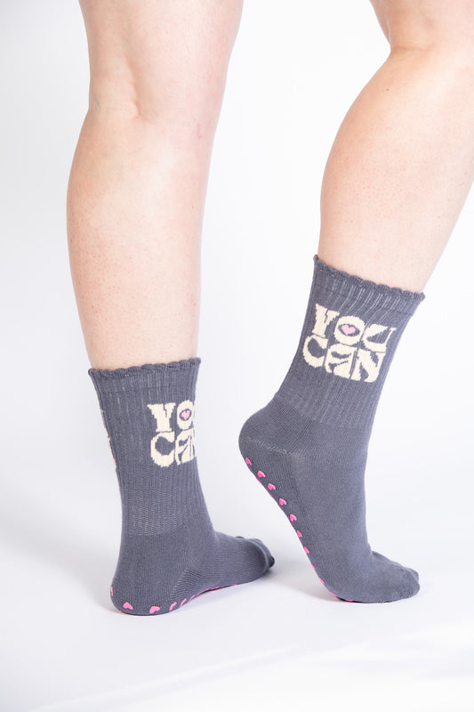 Model wearing charcoal gray Pilates grip socks with pink heart-shaped grips and “YOU CAN” text in vintage varsity font.