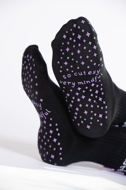 Close-up of lavender grip pattern and “so cutesy very mindful” text on soles of black Pilates socks.