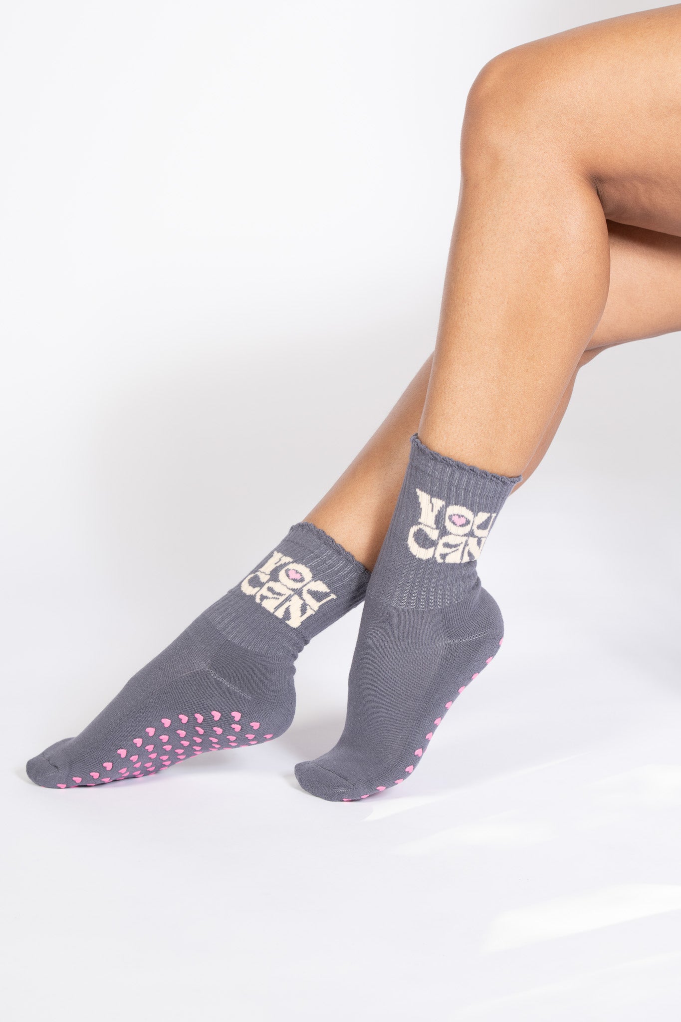 Model sitting cross-legged in gray grip socks featuring pink heart grips and affirming “YOU CAN” ankle text.