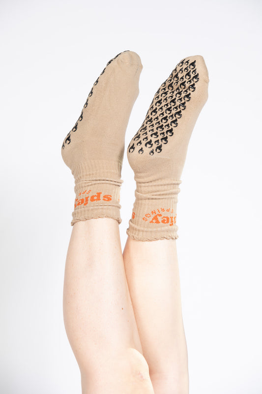 Model wearing tan Pilates grip socks with black flame grips and “spicy springs” text in orange.